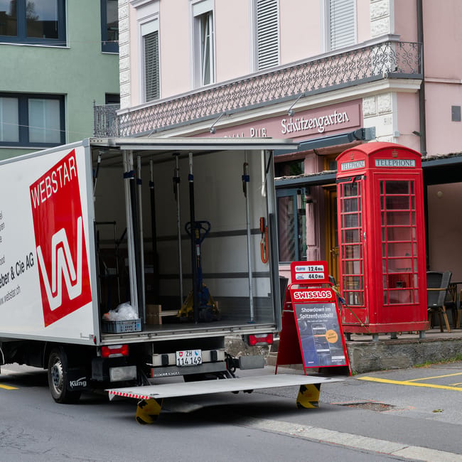 A manwithvanworcesterpark.co.uk moving van with an open rear compartment parked on the street in front of a building with a pink and white facade and a decorative railing balcony. The van’s loading area contains a few tools, including a blue hand truck and a plastic bag. It is positioned on a mobile platform with wheel guides, ready for a home relocation or furniture transport. Nearby, a traditional red British telephone box and a small blackboard sign advertising Swiss rolls are visible, placed on the sidewalk next to the building’s entrance. The scene depicts an urban environment with the van prepared for packing and moving services, highlighting the logistical process of loading household items onto the vehicle before transport to a new property. Natural lighting and clear weather suggest an outdoor loading process typical of a house removal operation, supported by the presence of equipment and materials used for packing and transportation.