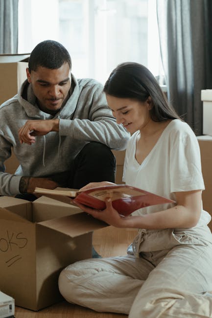 A young man and woman sit on a wooden floor inside a well-lit living room with a large window and curtains. The woman, dressed in casual light-colored clothing, is holding an open red book, while the 