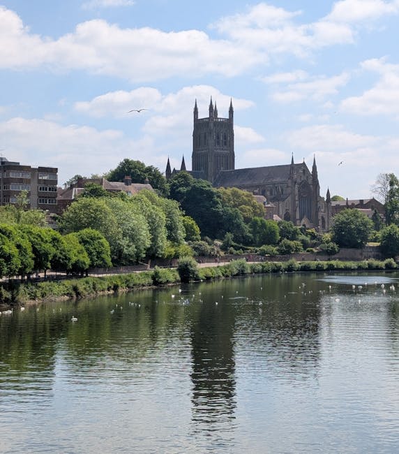 A view of a calm river with gentle ripples reflecting the blue sky and scattered clouds above. On the far side of the water, there is a line of lush green trees and bushes lining the riverbank. Behind the trees, a historic stone church with tall, pointed spires and gothic architecture dominates the skyline, set against neighboring residential buildings. In the foreground, minimal outdoor furniture or objects are visible, with no furniture or boxes related to house removals. The scene appears to be taken from a paved walkway or pavement area adjacent to the river, under natural daylight. Although the image primarily features a scenic landscape, it provides context for the local environment relevant to house removals and moving logistics, aligning subtly with relocation services such as those offered by Man with Van Worcester Park.