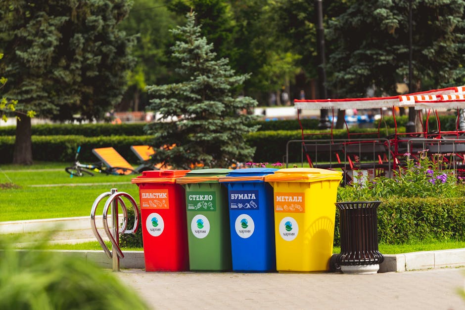 Four brightly coloured waste bins arranged in a row on a paved area outside a property, each designated for different types of waste: red for plastic, green for glass, blue for paper, and yellow for metal. The bins are positioned next to a black plastic trash can, with a set of metal bike racks nearby, and are set against a backdrop of greenery including trees, bushes, and a lawn. In the background, there are outdoor seating areas with orange and black loungers, and some red outdoor furniture with a red-and-white striped canopy or barrier. The scene is well-lit with natural daylight, illustrating a typical outdoor waste disposal setup during a home removal or moving process, as part of a house clearance or bulky waste disposal service provided by Man with Van Worcester Park.