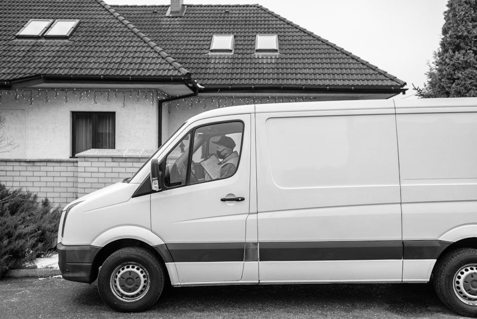 A man dressed in a red and black jacket, blue beanie, and black trousers stands with arms crossed next to a light grey commercial van with its rear doors open, revealing the empty cargo area lined wit
