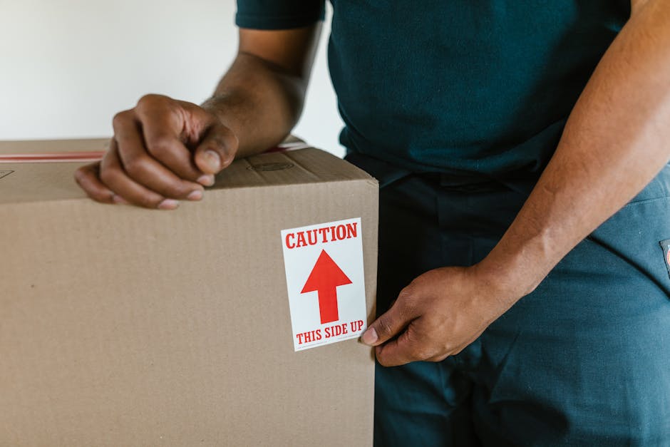 Close-up of a man wearing a dark green shirt and blue trousers, standing inside a property near a doorway, preparing to load a cardboard box onto a moving van. The box is sealed with packing tape and has a red and white caution sticker with an upward arrow and the text 'Caution, This Side Up.' The setting suggests a home relocation or furniture transport process, with the man handling the box as part of packing and moving activities associated with house removals or local moving services. The background is minimal and the lighting is natural, focusing on the careful handling of the packed items to ensure safe transport. The image reflects professional packing and loading procedures used by companies like Man with Van Worcester Park, aligned with service categories such as removals, and referencing the importance of proper handling when relocating belongings.