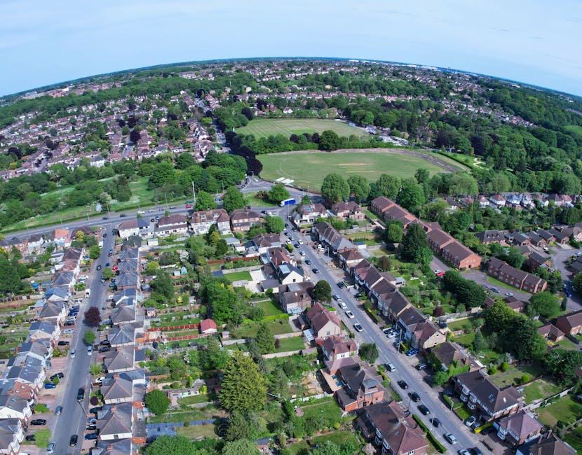 An aerial view of a riverside town featuring a mix of residential and commercial buildings with pitched roofs and brick facades, situated along a waterway with several boats docked at the quay. The st