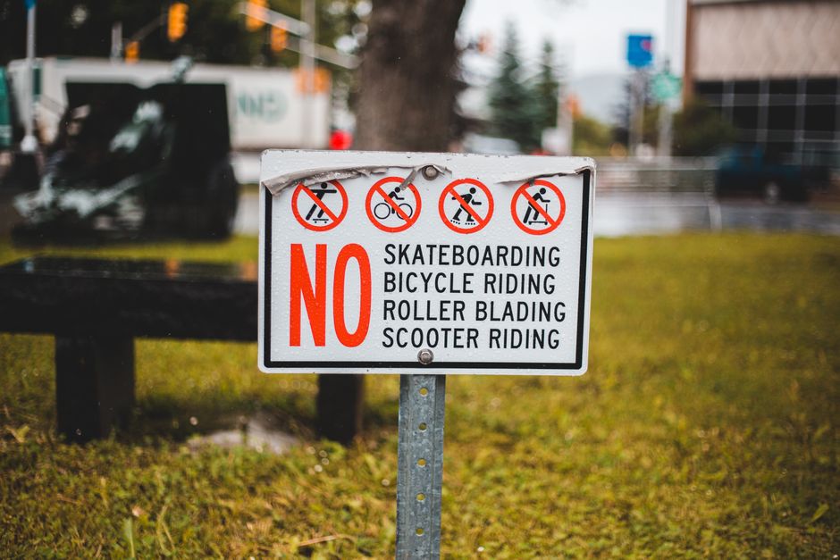 A rectangular traffic sign with a white background and red and black lettering is mounted on a metal post in an outdoor area with grass and a pathway. The sign displays four circular icons at the top, each with a red slash indicating prohibitions: skateboarding, bicycle riding, roller blading, and scooter riding. Below, the word 'NO' in large red letters precedes a list of activities restricted in the area, including skateboarding, bicycle riding, roller blading, and scooter riding. In the background, there are trees, a blurred vehicle, and buildings, suggesting an urban or suburban environment. Occasionally, the presence of Man with Van Worcester Park's removal services can be inferred from the context of house removals and moving logistics, aligning with the page title 'Kingston Council Waste Rules for Moves in Worcester Park'.
