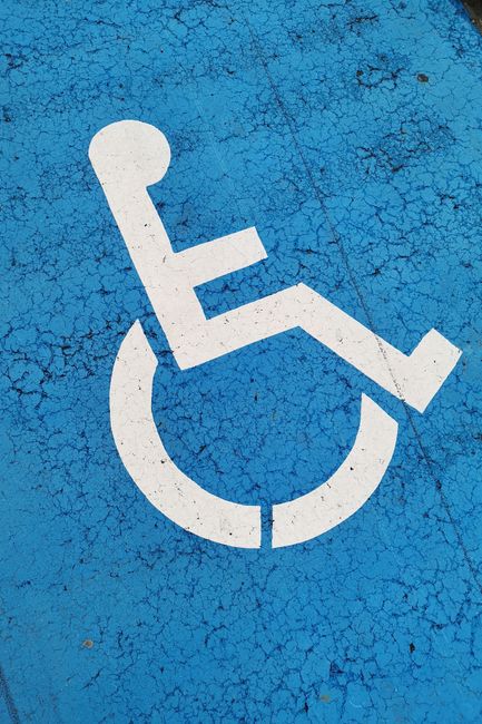 Close-up view of a blue disabled parking space surface with a white wheelchair symbol painted on it. The paint shows signs of cracking and weathering. In the background, there is a section of a pavement or driveway, indicating an accessible parking area typically used for home relocation or property transfers. The image is taken outdoors with natural lighting, highlighting the condition of the markings and surface. This visual is relevant for content about parking regulations and logistics involved in house removals and furniture transport, as handled by Man with Van Worcester Park.