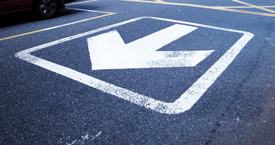A painted white arrow on a dark asphalt pavement indicating a direction, positioned within a rectangular border. The surrounding area features parking lines and a parked vehicle partially visible at the top edge of the image. The scene is outdoors, with natural lighting that highlights the texture of the asphalt and the freshly painted markings. This type of pavement marking is commonly found in parking lots to guide vehicle movement during house removals or moving processes. The presence of such markings suggests the location is an accessible loading or unloading zone, which relates to the moving logistics managed by Man with Van Worcester Park, specialising in removals services and home relocation support. The clean and clear directional arrow helps facilitate an efficient loading and unloading process, ensuring safety and compliance with parking and transport regulations for Worcester Park.