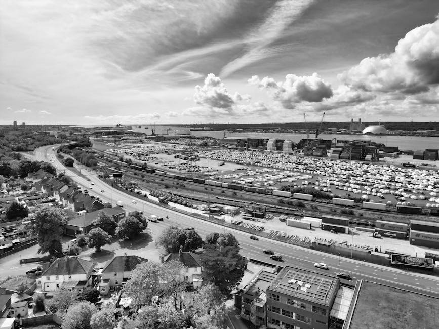 A black-and-white aerial view of an urban industrial area with a large shipping yard filled with stacked cargo containers, cranes, and trucks. In the foreground, a residential neighbourhood with several houses surrounded by trees is visible, along with roads and vehicles. Further back, a body of water with a bridge crossing over it is seen, with a distinctive domed building in the distance under a partly cloudy sky. This image, associated with house removals and relocation services, illustrates the scale and logistics involved in furniture transport and loading processes, relevant to professional removals in Worcester Park as provided by Man with Van Worcester Park.
