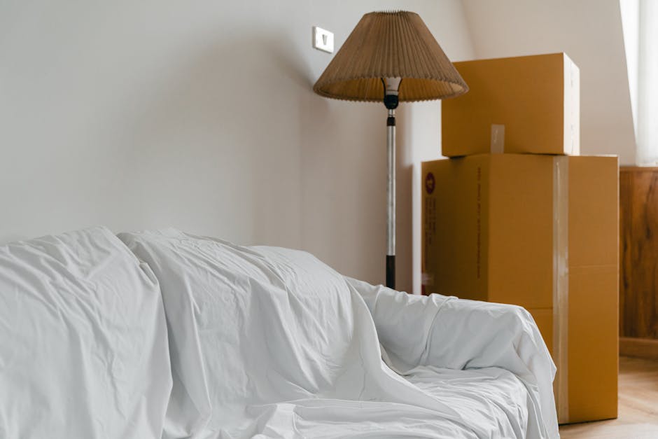 Interior view of a room during a home relocation process, featuring a white sofa covered with protective fabric on the left side of the image. Behind the sofa, there is a tall, vintage-style floor lamp with a pleated beige lampshade casting subtle light. To the right of the lamp, three cardboard boxes of varying sizes are stacked, with the smallest on top. The boxes are sealed with clear packing tape and appear to be used for packing household items. The setting includes a wooden sideboard or cabinet partially visible on the far right and a window partially seen in the background, allowing natural light to illuminate the scene. The environment indicates a typical living room, with furniture and packing materials organized in preparation for a furniture transport or moving process. The image reflects the packing and loading phase often associated with professional removals services by Man with Van Worcester Park, emphasizing careful handling of household belongings during an urgent same-day move in Worcester Park.