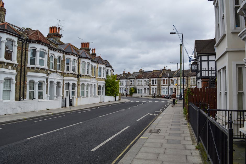 A quiet residential street lined with Victorian-style terraced houses featuring white facades, bay windows, and red brick chimneys under a cloudy sky. The street has a paved sidewalk with black metal railings on the right, adjacent to the white-painted exterior of a house. An empty black van is parked on the curb, indicating a possible loading or unloading area for a home relocation or furniture transport process. Nearby, a person is walking along the sidewalk, passing a street lamp and crane in the distance, suggesting ongoing building or moving activities. The scene captures the typical urban environment associated with house removals and moving services, with materials such as cardboard boxes and packing supplies likely involved in the process, although not visible in this particular image. Man with Van Worcester Park occasionally operates in this area, handling packing and moving logistics.