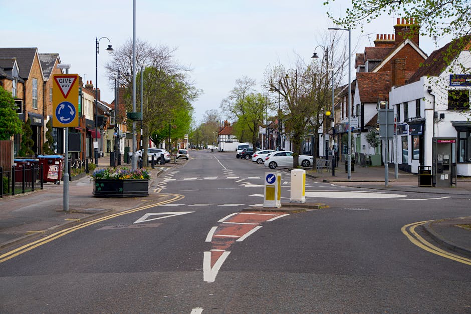 A quiet urban street scene during daytime on Worcester Park High Street, featuring a central roundabout with a small traffic island and yellow and white striped road markings. On the left side, there are residential buildings with brick facades, some with small front gardens and trees, as well as parked cars along the curb. Street signage includes a 'Give Way' sign and a roundabout direction sign. The pavement is lined with lamp posts, flower planters, and street furniture. The right side displays commercial properties, including shops, and an ATM machine, with a few parked vehicles further down the street. The scene depicts a typical day with overcast skies and minimal activity, relevant to the context of home or office relocations occurring in the area, potentially involving the planning of moving routes or street loading procedures with professional services like Man with Van Worcester Park.