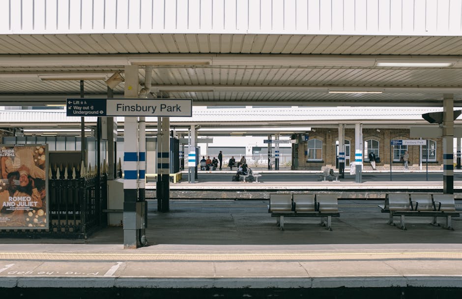 The platform at Finsbury Park railway station in daylight, showing several people seated on benches and walking along the station’s waiting area. The scene includes the station sign, which reads 'Finsbury Park,' and a poster advertising 'Romeo and Juliet' mounted on a metal fence near the platform entrance. The platform surface is paved with concrete, and there are multiple metal benches placed along the waiting area. The station is partially sheltered by a roof with visible lighting fixtures, supported by metal poles painted with blue and white stripes. In the background, across the tracks, there are additional platforms, waiting areas with more people, and railway buildings constructed from brick and glass. The environment appears calm with ample natural light, and there are no visible moving objects or items related to house removals, packing, or furniture transport within the scene. This image depicts a typical train station setting, illustrating the infrastructure encountered during house relocations or transport logistics handled by Man with Van Worcester Park.
