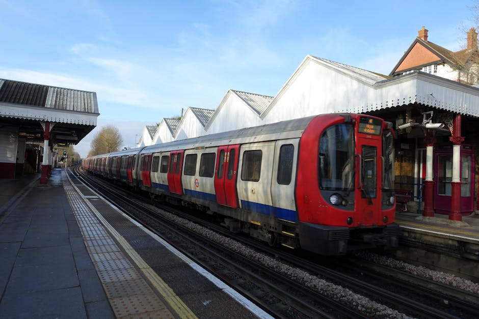 A modern London underground train with a red, white, and blue exterior parked at Worcester Park station platform during daytime. The train is on the left side of the image, with its doors closed, alongside a platform made of concrete with tactile paving strips near the edge for accessibility. To the right, the station building features white wooden gabled roofs with scalloped trim and deep red decorative columns supporting the platform roof. Several cardboard boxes wrapped in plastic are visible on the platform, some being carried or prepared for loading. Inside the station, there are luggage trolleys and transportation straps, indicating a home relocation or furniture transport process managed by Man with Van Worcester Park. The background shows a clear sky, trees, and residential houses with brick chimneys, suggesting a suburban setting. The image captures the typical scene during a professional moving service involving loading and unloading at a train station.